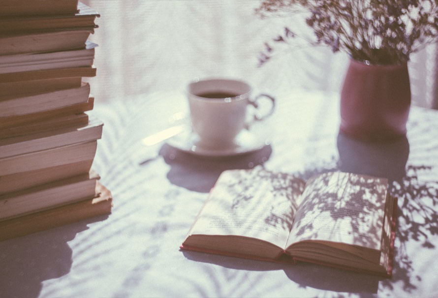 Stack of books beside an open book, a coffee cup, and dried flowers on a sunlit table